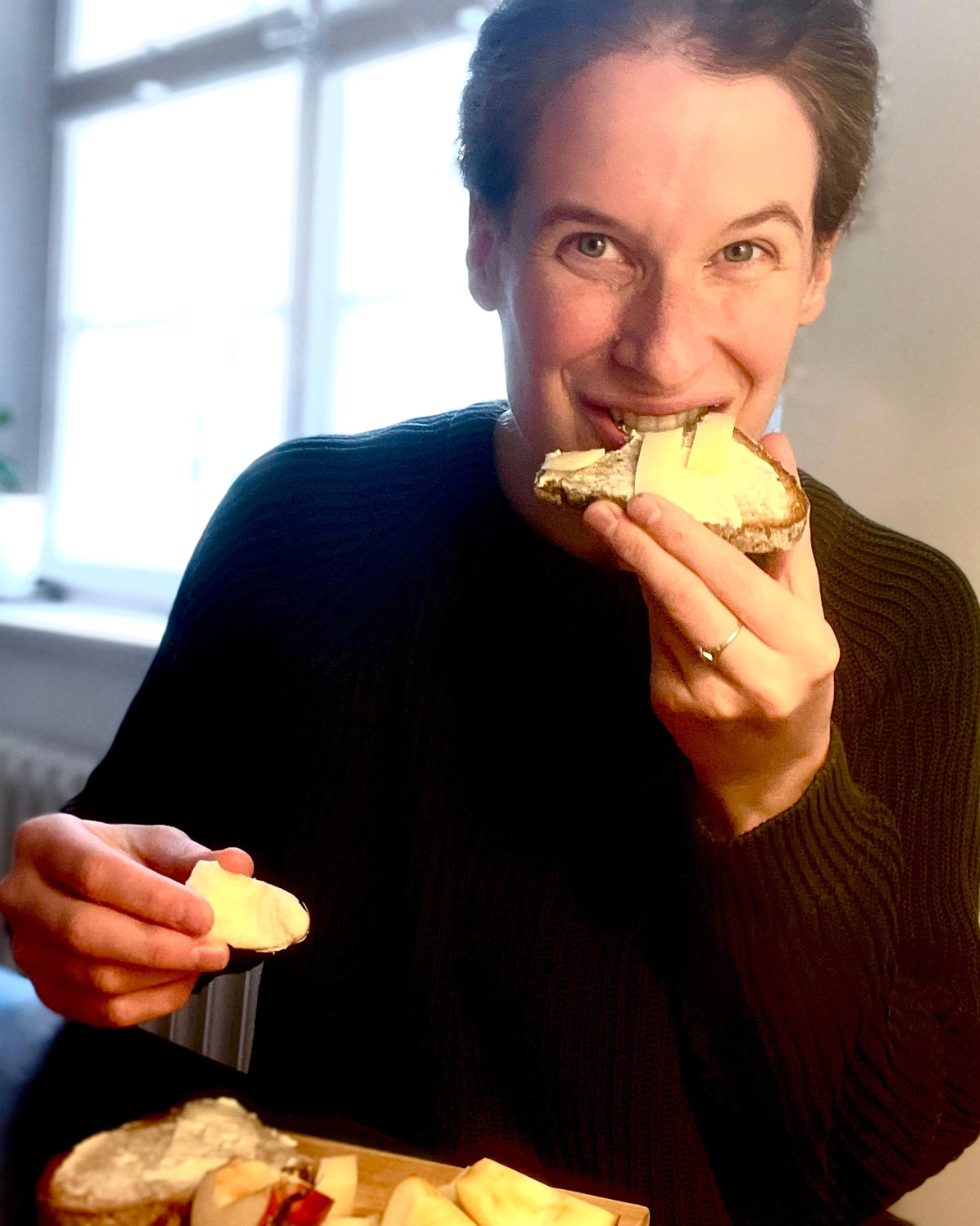 Marie Luisa Kerkhoff sitzt an einem Tisch vor einem Fenster und beißt lachend in ein Käsebrot. Auf dem Tisch liegt ein Holzbrett mit weiteren Brotscheiben und Apfelstücken. Sie trägt einen dunklen Strickpullover.