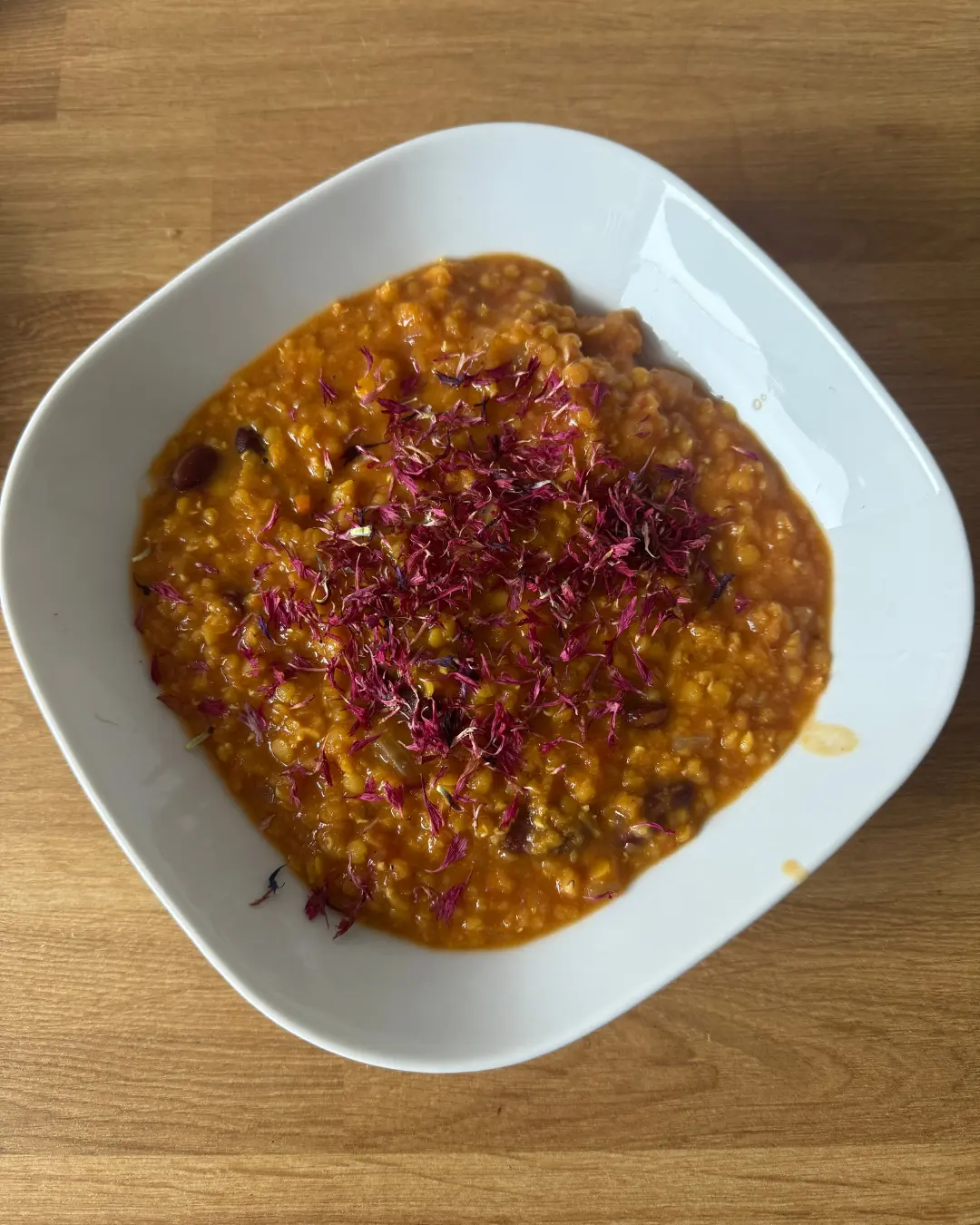 In einer weißen, quadratischen Schale liegt ein kräftig orangefarbenes Linsencurry (Dal), angerichtet mit roten Blütenblättern. Die Schale steht auf einem hellen Holztisch.