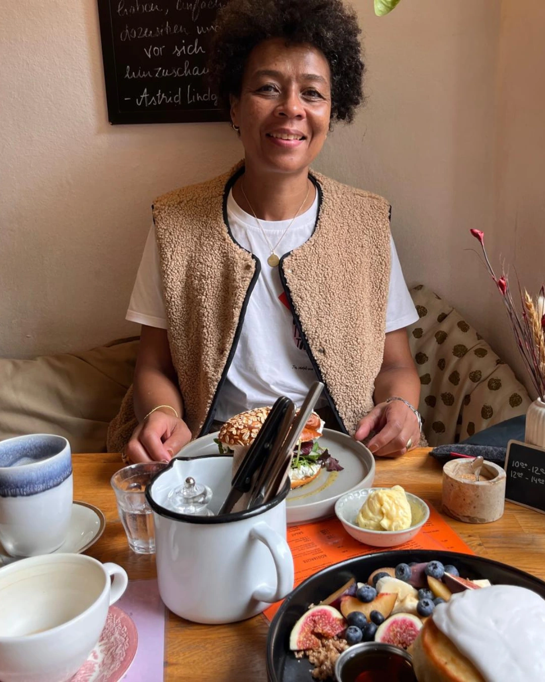 Susanne Amar in einem Café sitzend, auf dem Tisch stehen ein Bagel mit Avocado, Frischkäse und Rührei auf einem Teller, davor Kaffee, Wasser und ein Teller mit Obst.