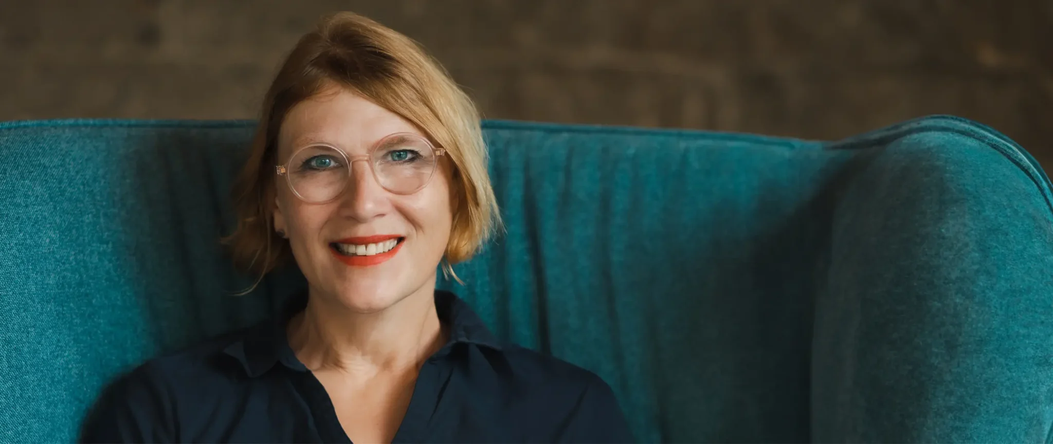 Christa Fajen sitzt auf einem petrolfarbenen Sessel. Sie trägt eine dunkelblaue Bluse, hat blonde halblange Haare und eine transparente runde Brille. Sie lächelt direkt in die Kamera.